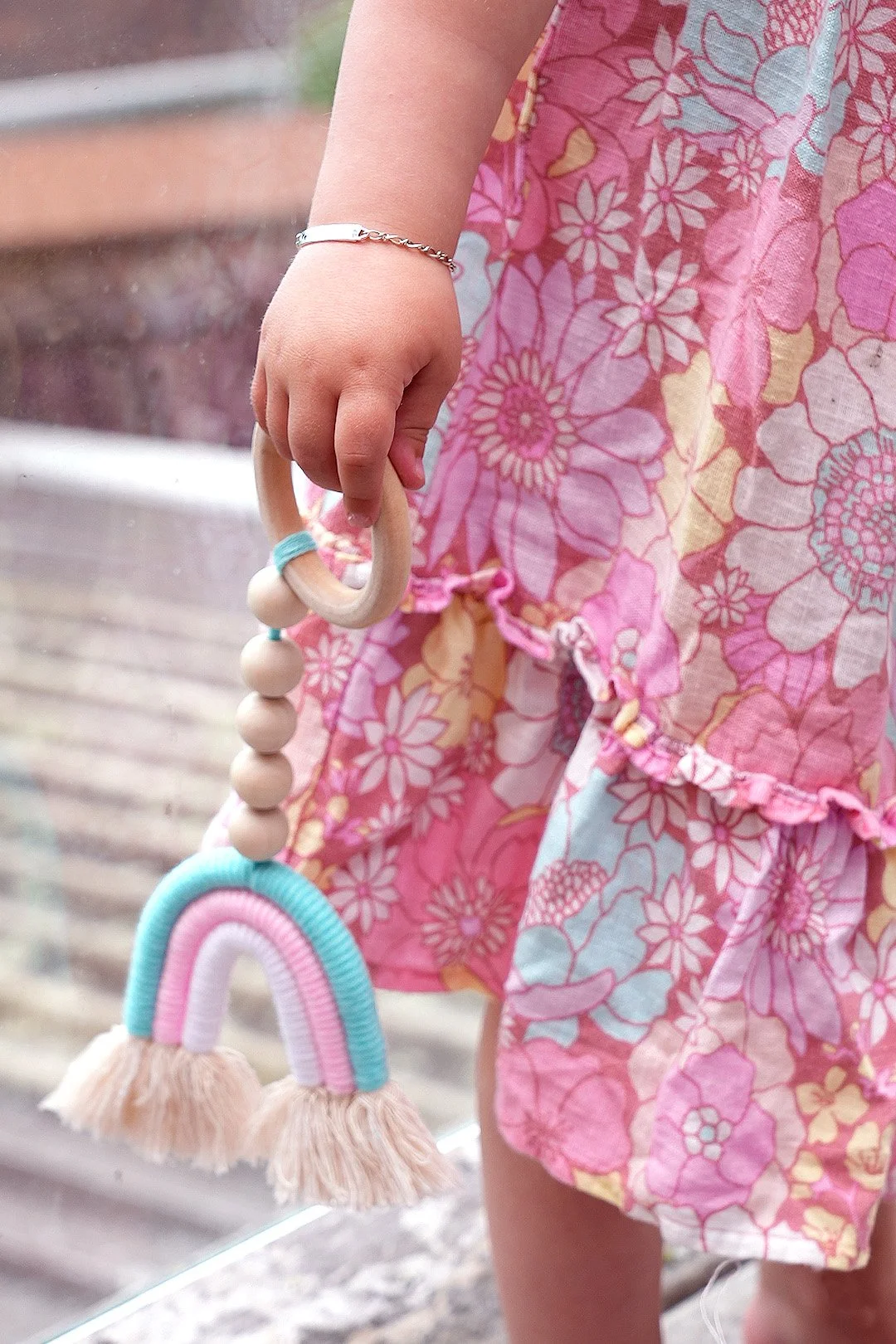 A young girl is holding a rainbow-shaped teething toy with beads and tassels, wearing a pink floral dress.