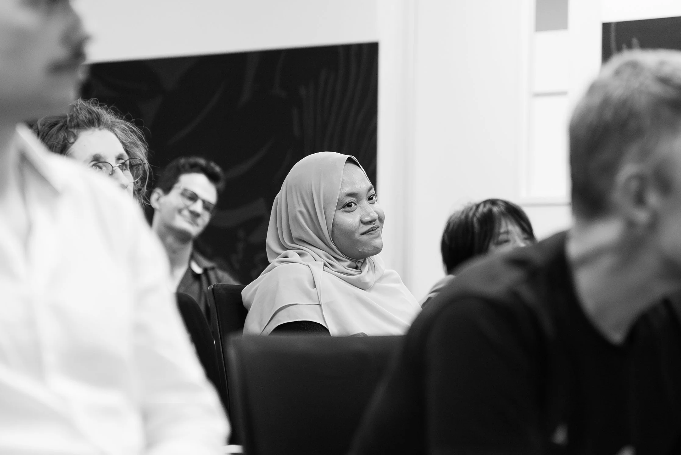 A diverse group of people sitting attentively at a meeting or presentation, with some smiling and listening.