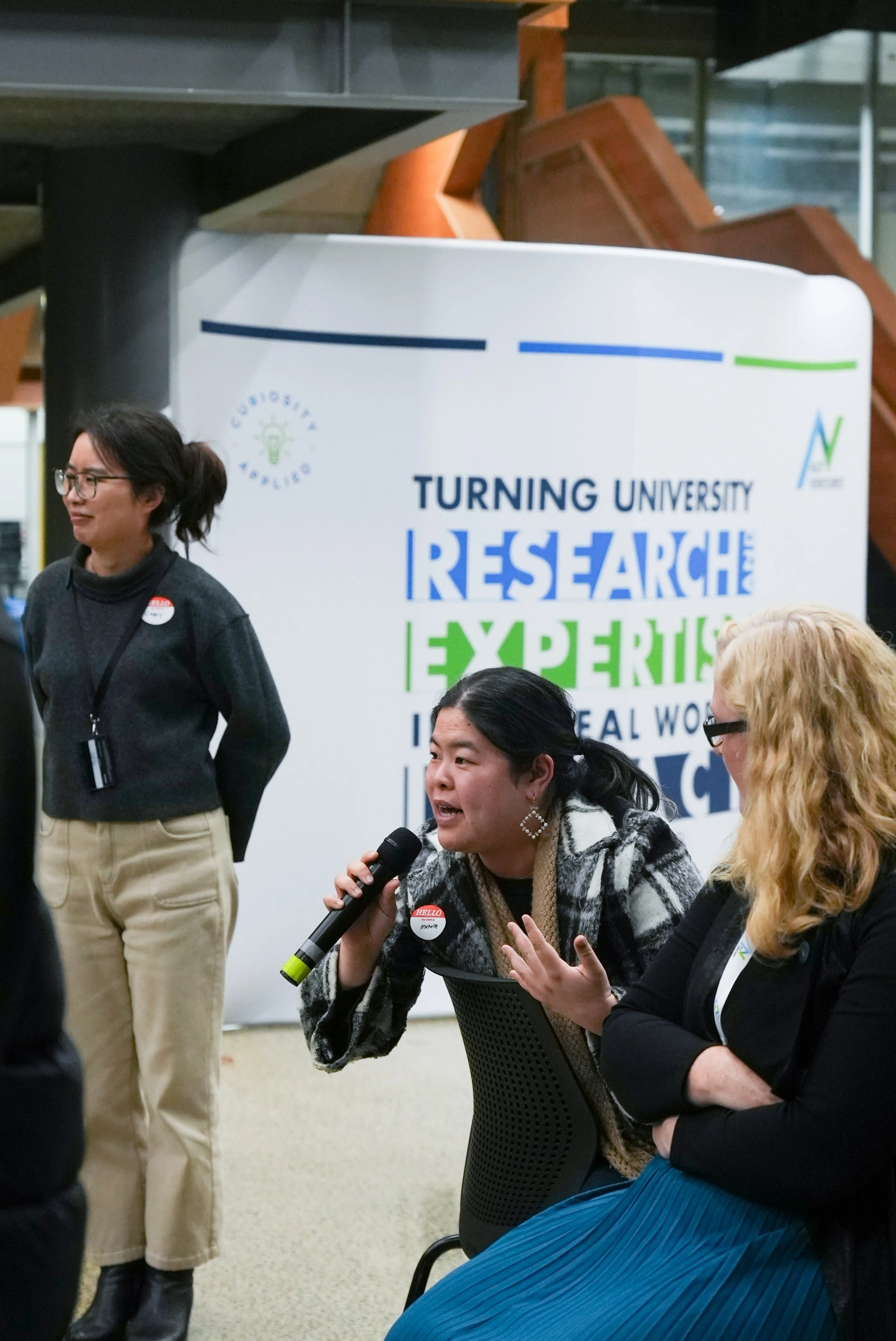 A woman speaking into a microphone at a research event with a Turning University sign in the background, three women are visible, two of whom are seated and one standing, all engaged in conversation.