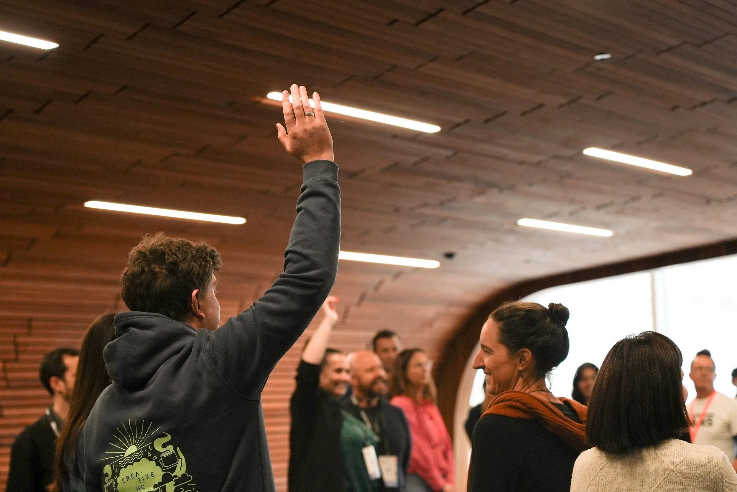 A person with curly hair wearing a black hoodie with a green graphic is raising their hand, engaging with a group of smiling people in an indoor setting with a wooden ceiling.
