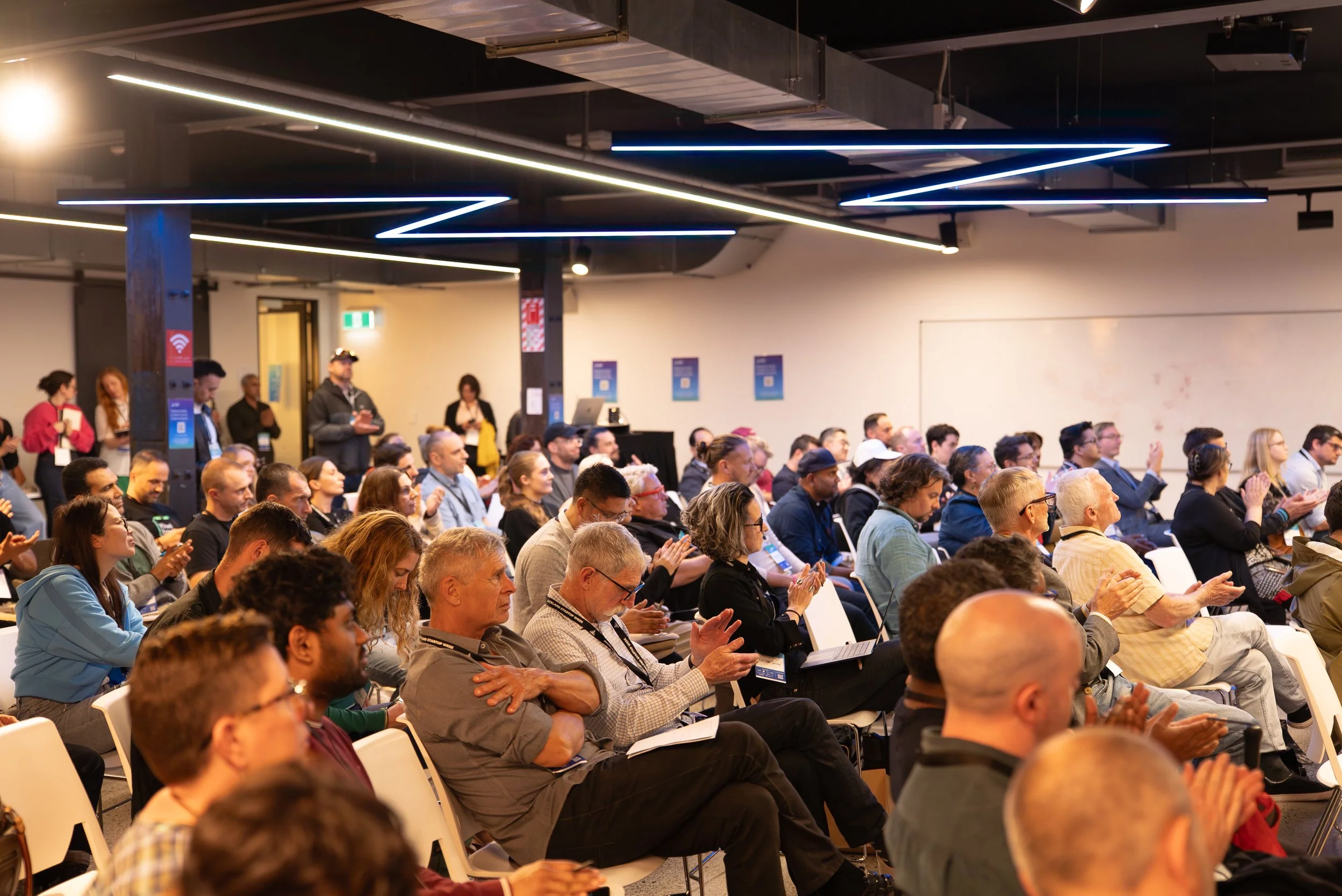 Crowd of people attending a conference, seated and clapping in a modern indoor venue with geometric lighting.