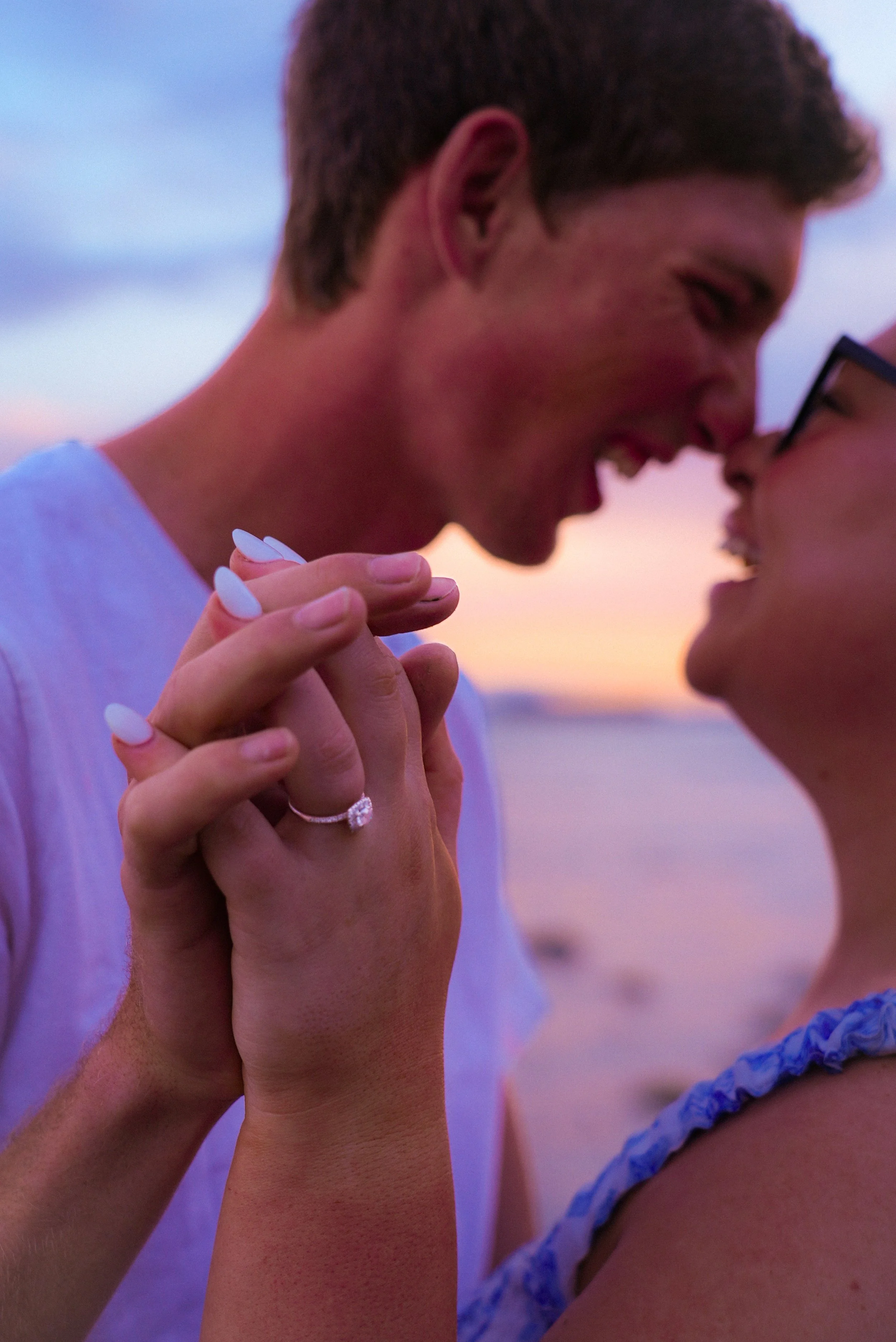A couple holding hands and smiling at each other on the beach during sunset, with the woman wearing an engagement ring.
