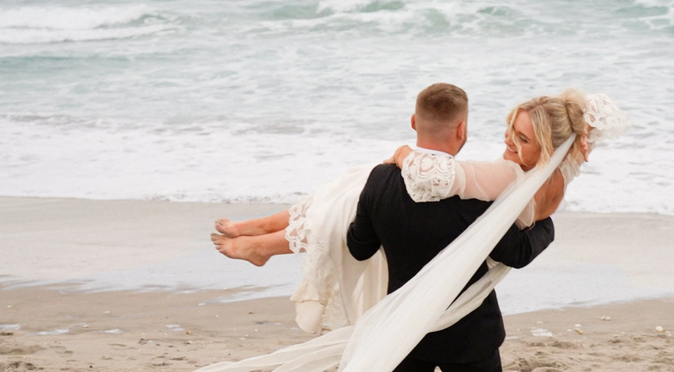A couple on the beach, with the man carrying the woman in his arms, by the ocean waves. The woman is wearing a white dress and the man a dark suit.