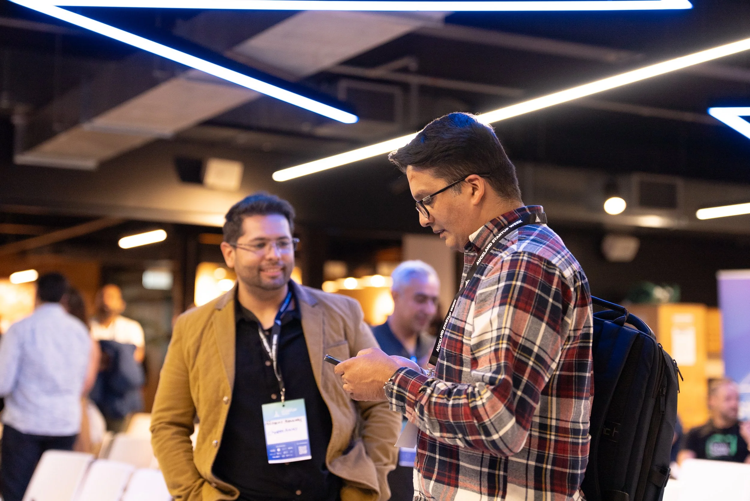 Two men talking at a conference, one is showing something on his phone to the other. The man on the left is wearing glasses and a brown blazer, the man on the right is wearing glasses, a plaid shirt, and has a backpack.