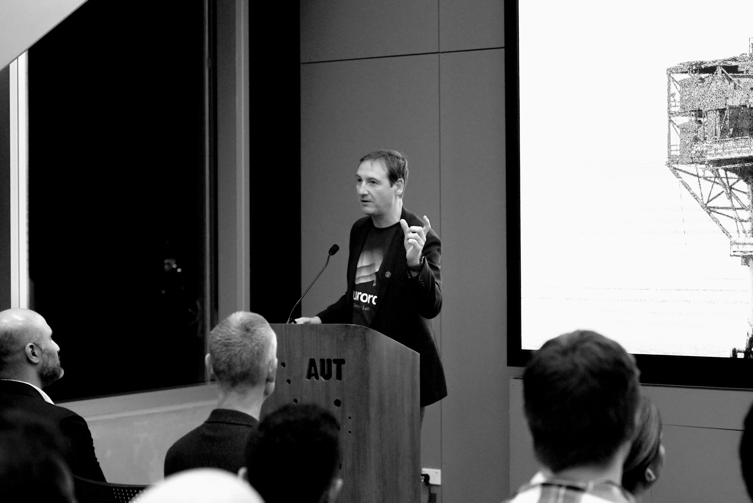 A man giving a presentation at a conference, standing behind a podium labeled 'AUT', with a large screen displaying an image of a structure to his right, audience members listening attentively.