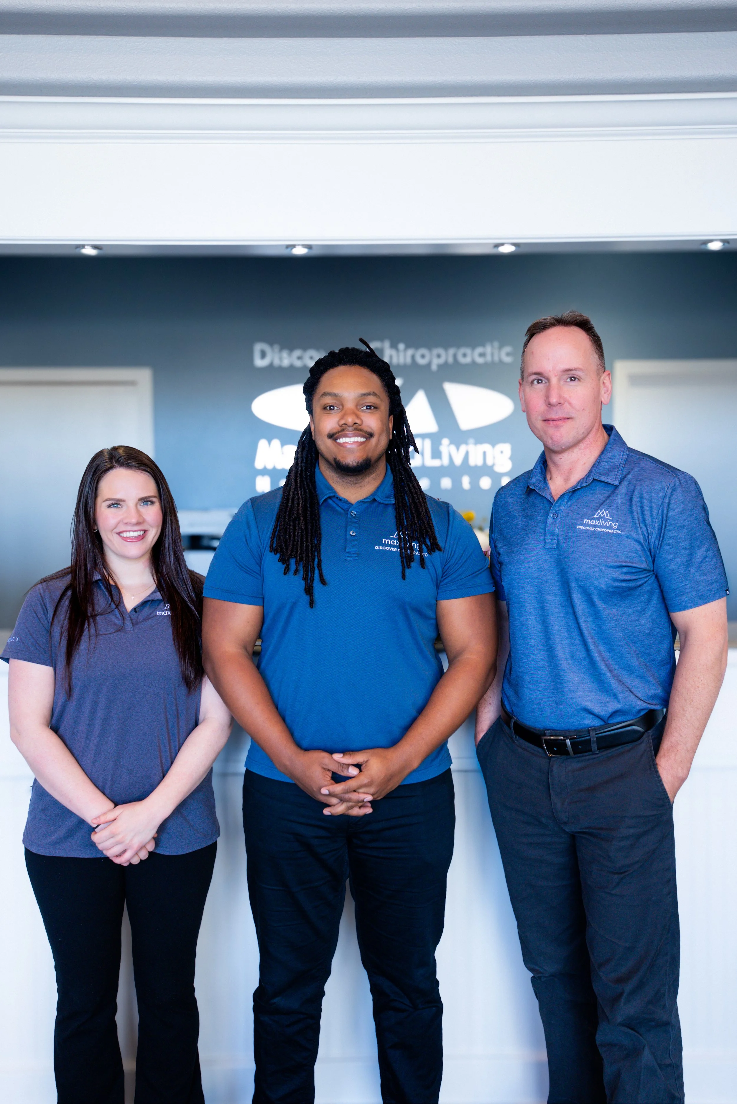 Three people standing in front of a sign that reads 'Discover Chiropractic MaxLiving' inside a wellness center, wearing blue and gray MaxLiving uniforms.