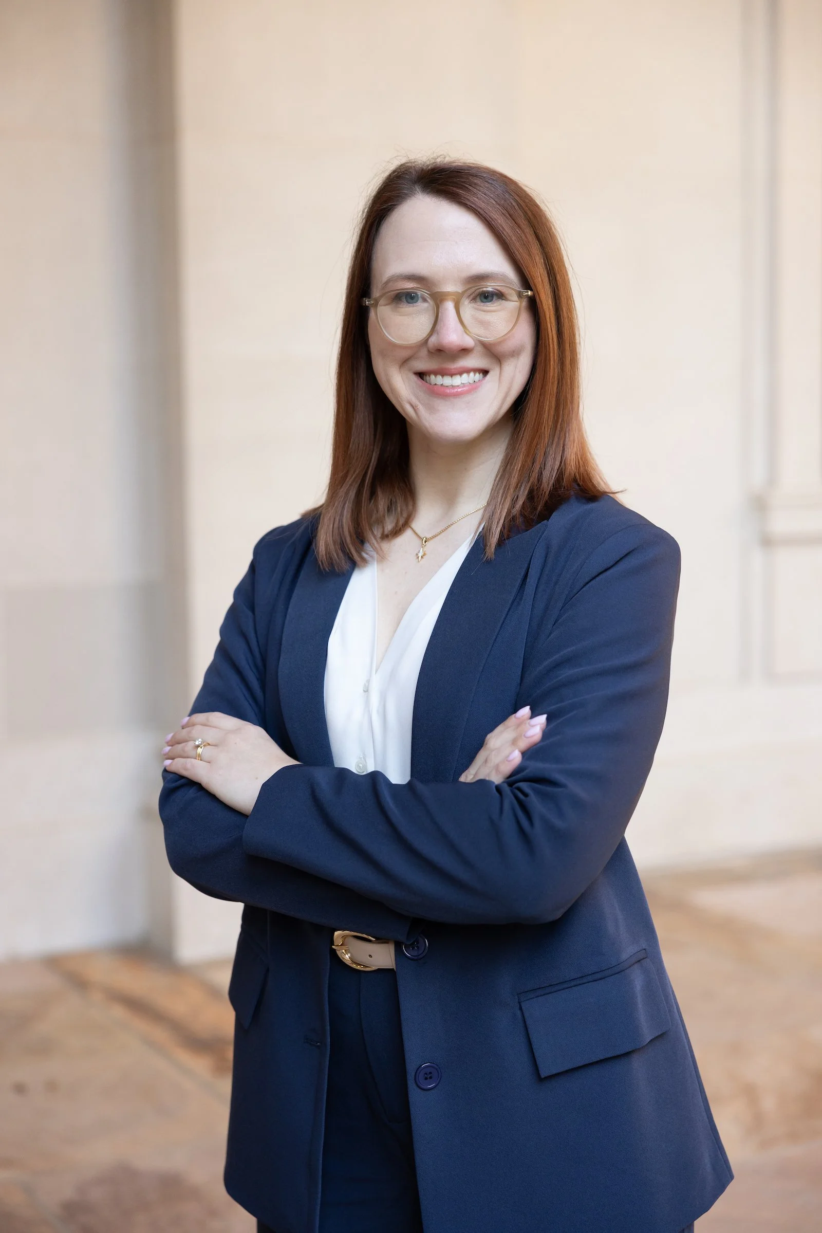 A professional young woman with red hair, wearing glasses, a navy blazer, and white shirt, standing with arms crossed in a light-colored room.