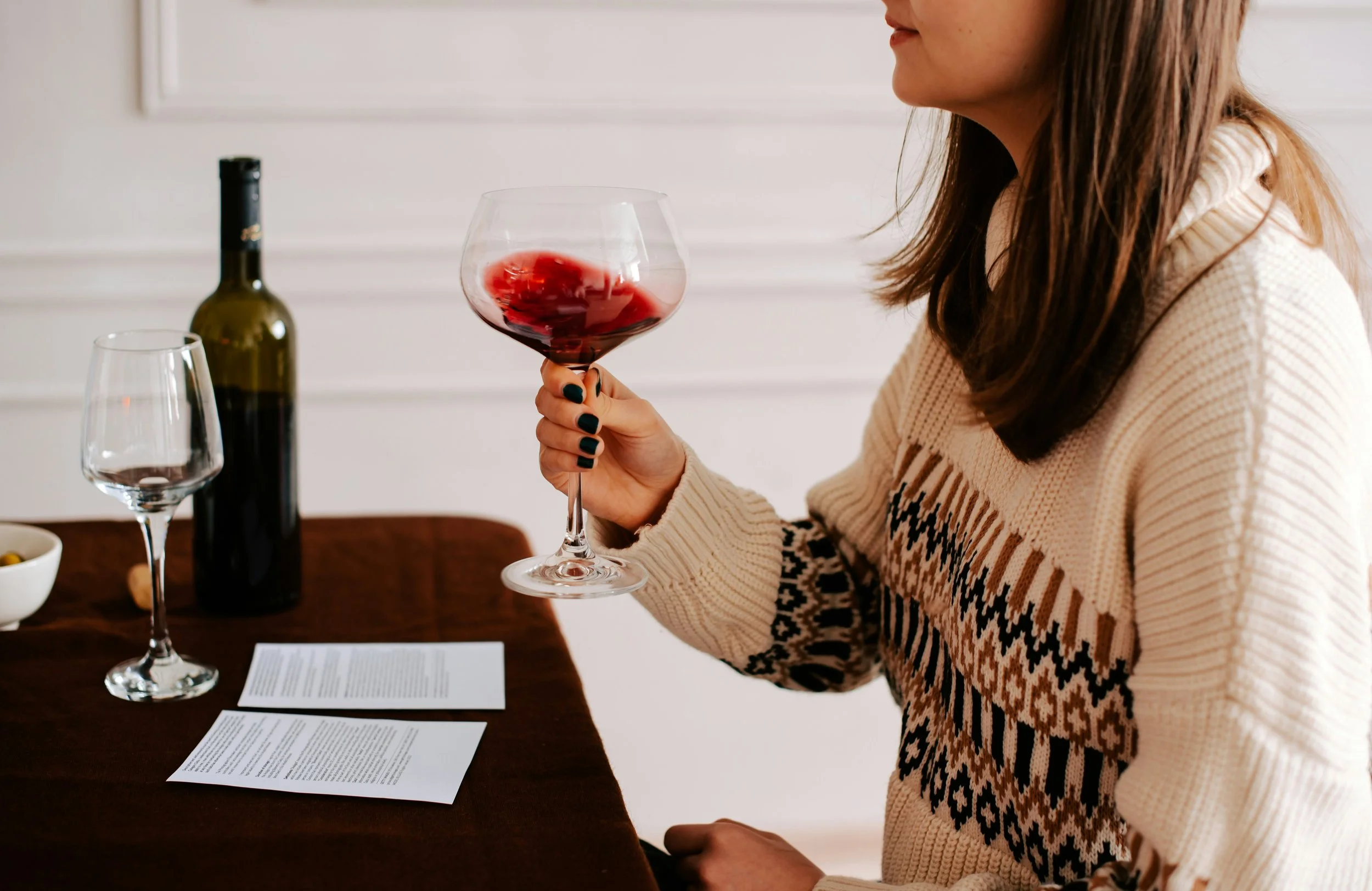 A brunette white woman in a cable knit sweater swirls a glass of red wine