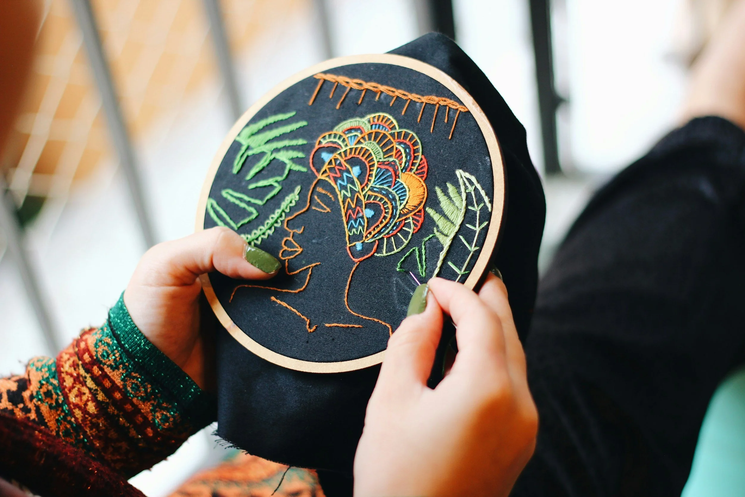 Personne brodant un motif coloré sur un tissu noir, montrant le profil d'un visage entouré de feuilles vertes et des motifs abstraits.