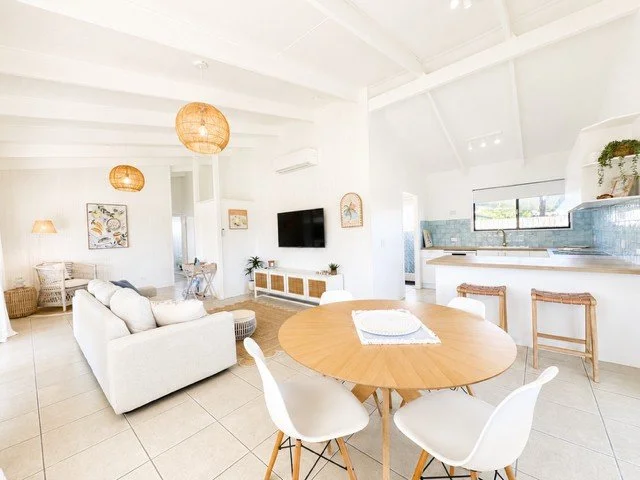 Bright, open-concept living and kitchen area with white walls and ceiling, tan tiled floor, a white sofa, wicker armchair, round wooden dining table with white chairs, and a blue-tiled kitchen counter with a window above, decorated with potted plants and wall art.