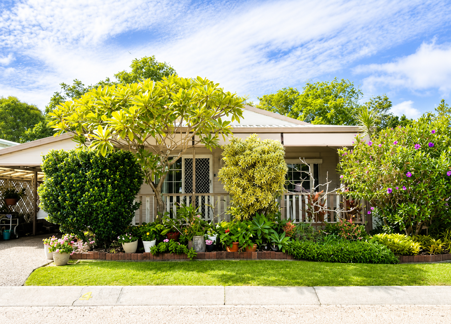 Front view of a house with a garden featuring various green plants and flowers, a white fence, and a bright blue sky with scattered clouds.