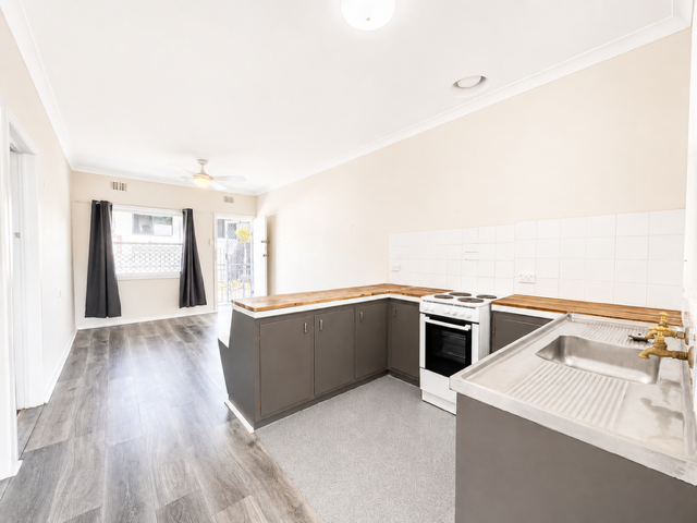 Empty kitchen and living area with gray cabinets, white stove, stainless steel sink, dark wood flooring, and a window with black curtains.