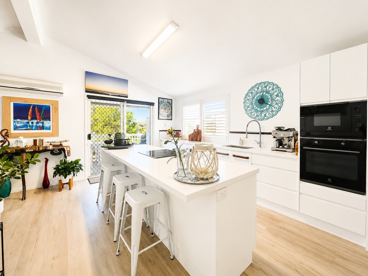 Bright, modern white kitchen with a central island, bar stools, and a window with blinds. Decor includes plants, artwork, and a decorative wall clock.
