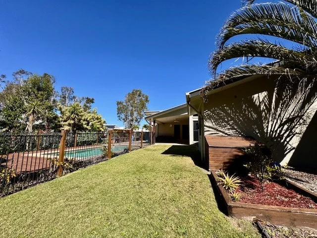 Backyard with a manicured grassy lawn, a swimming pool surrounded by a black metal fence, and a house with a porch, palm trees, and bushes under a clear blue sky.
