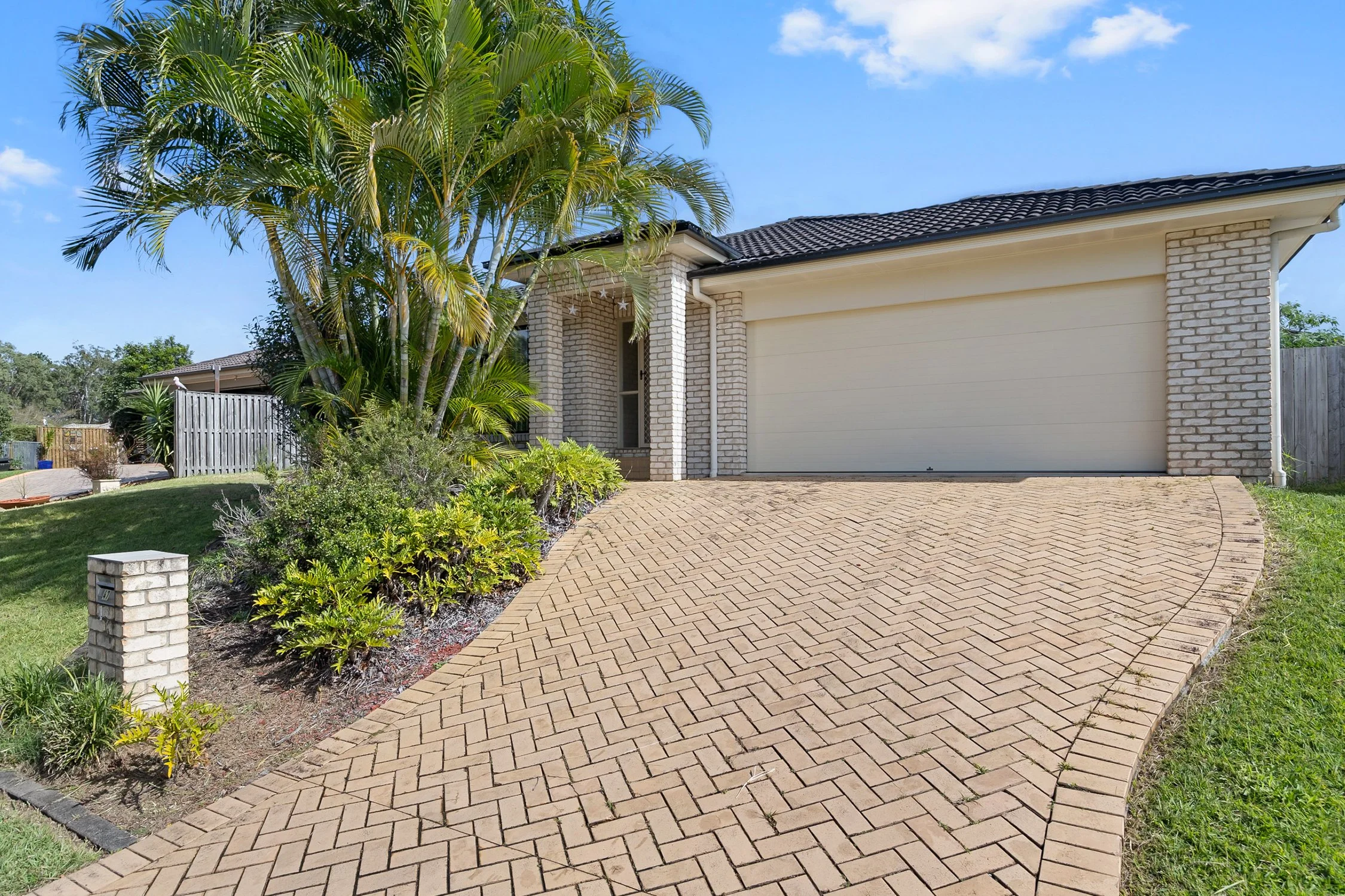 A house with a brick facade and a white garage door in a suburban neighborhood, with palm trees and a paved driveway in the front yard under a partly cloudy sky.