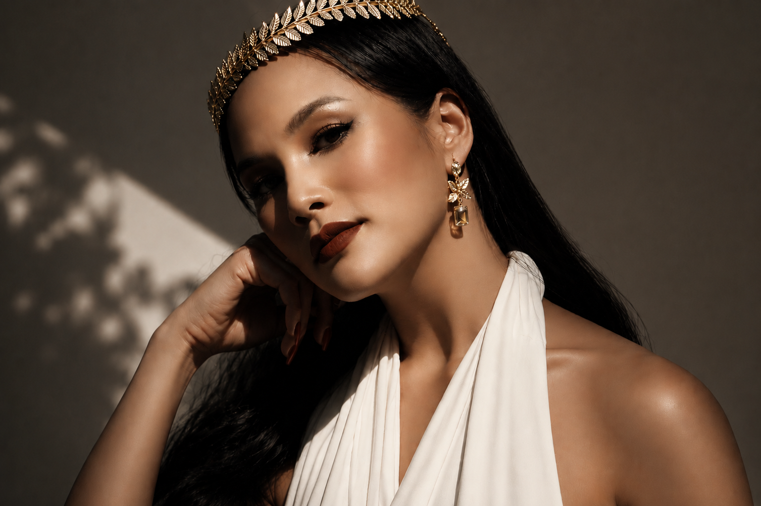 A woman with long dark hair, wearing a gold leaf headband and gold earrings, in a white dress with a pleated neckline, posing with her hand near her face against a neutral background with shadow patterns.
