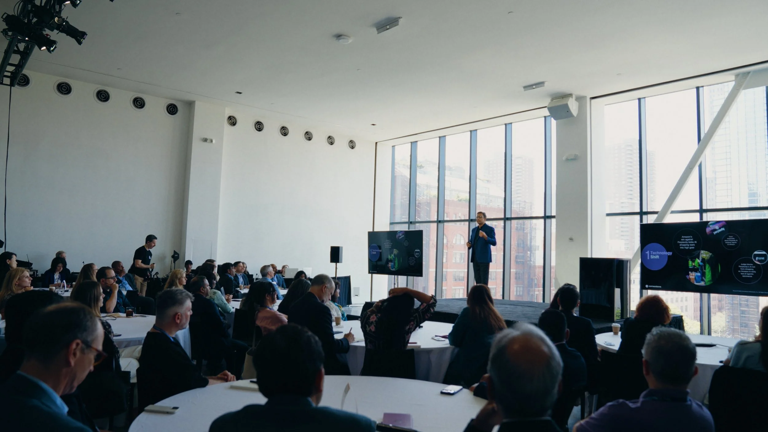 A speaker presenting at a conference with a large digital screen, in a bright, high-rise conference room filled with seated attendees.
