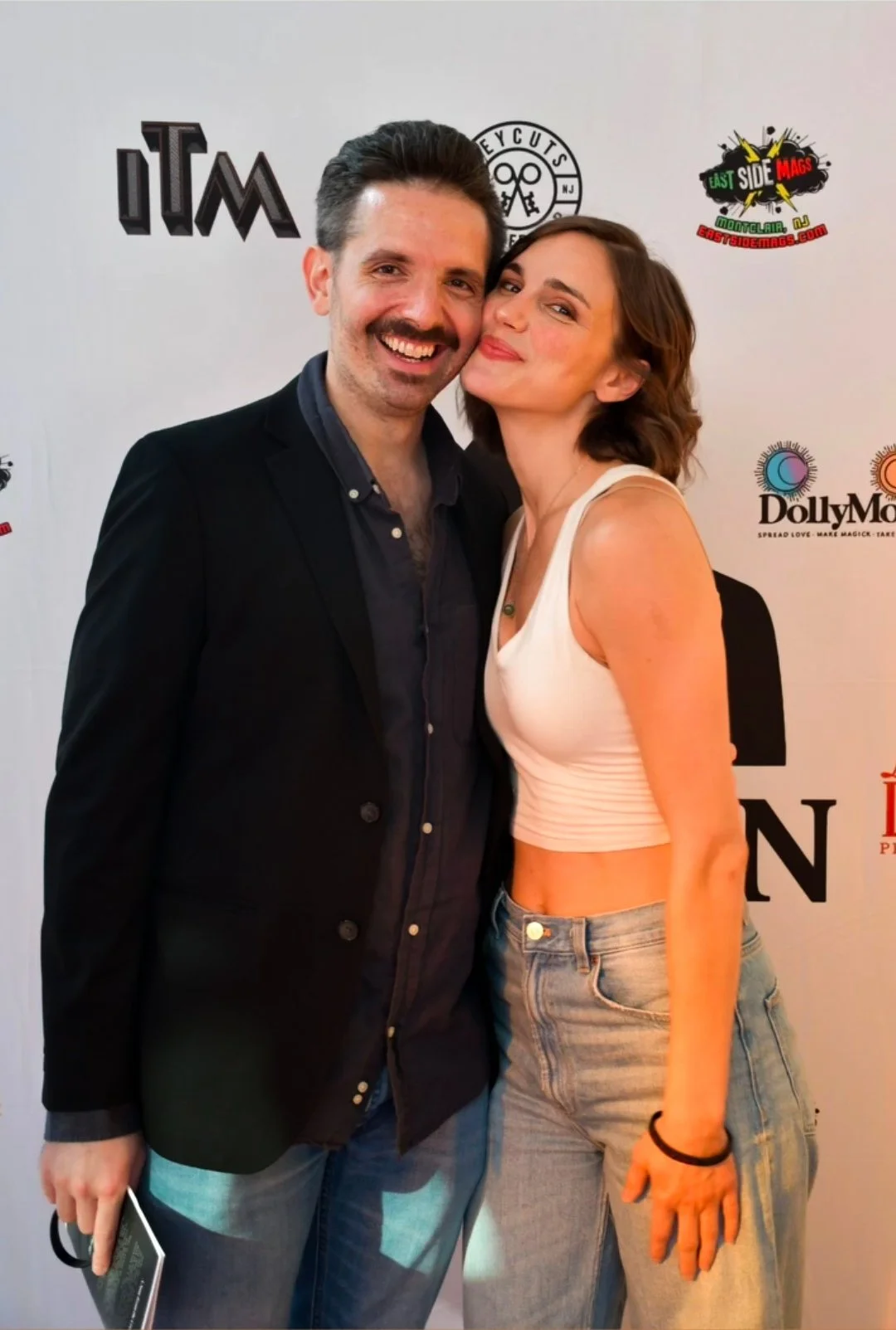 Two people posing for a photo at a promotional event, standing close and smiling. The man on the left has dark hair and a beard, wearing a dark blazer over a dark shirt. The woman on the right has short brown hair, wearing a white crop top and light blue jeans. They are in front of a backdrop with various logos.