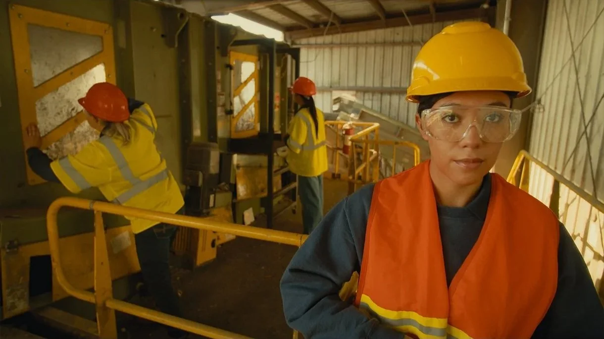 Three workers in safety gear working in an industrial setting, one in the foreground looking at the camera, and two in the background handling equipment.
