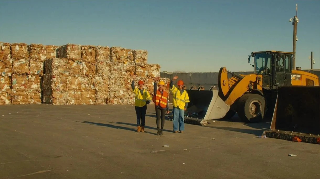 Three construction workers wearing safety vests and helmets stand near a large backhoe loader on a construction site, with a stack of bricks in the background.