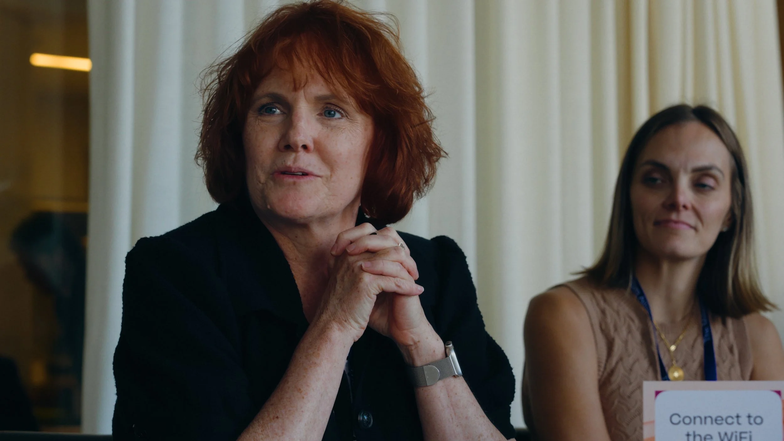 Two women sitting at a table during a conference or meeting. The woman in the foreground has red hair, is clasping her hands together, and appears to be speaking or listening. The woman in the background has brown hair, is smiling, and looking towards the other woman. A sign on the table says "Connect to the WiFi."