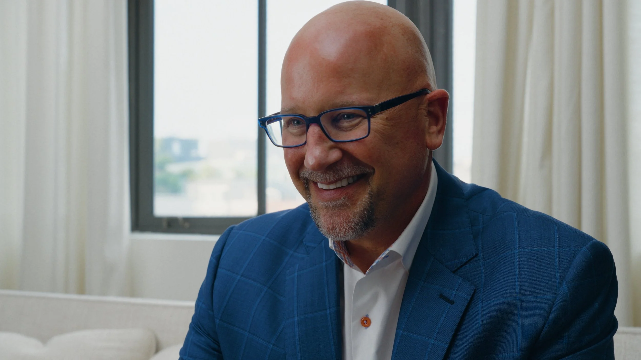 A bald man with glasses and a beard smiling, wearing a blue suit, sitting in a room with large windows and curtains.
