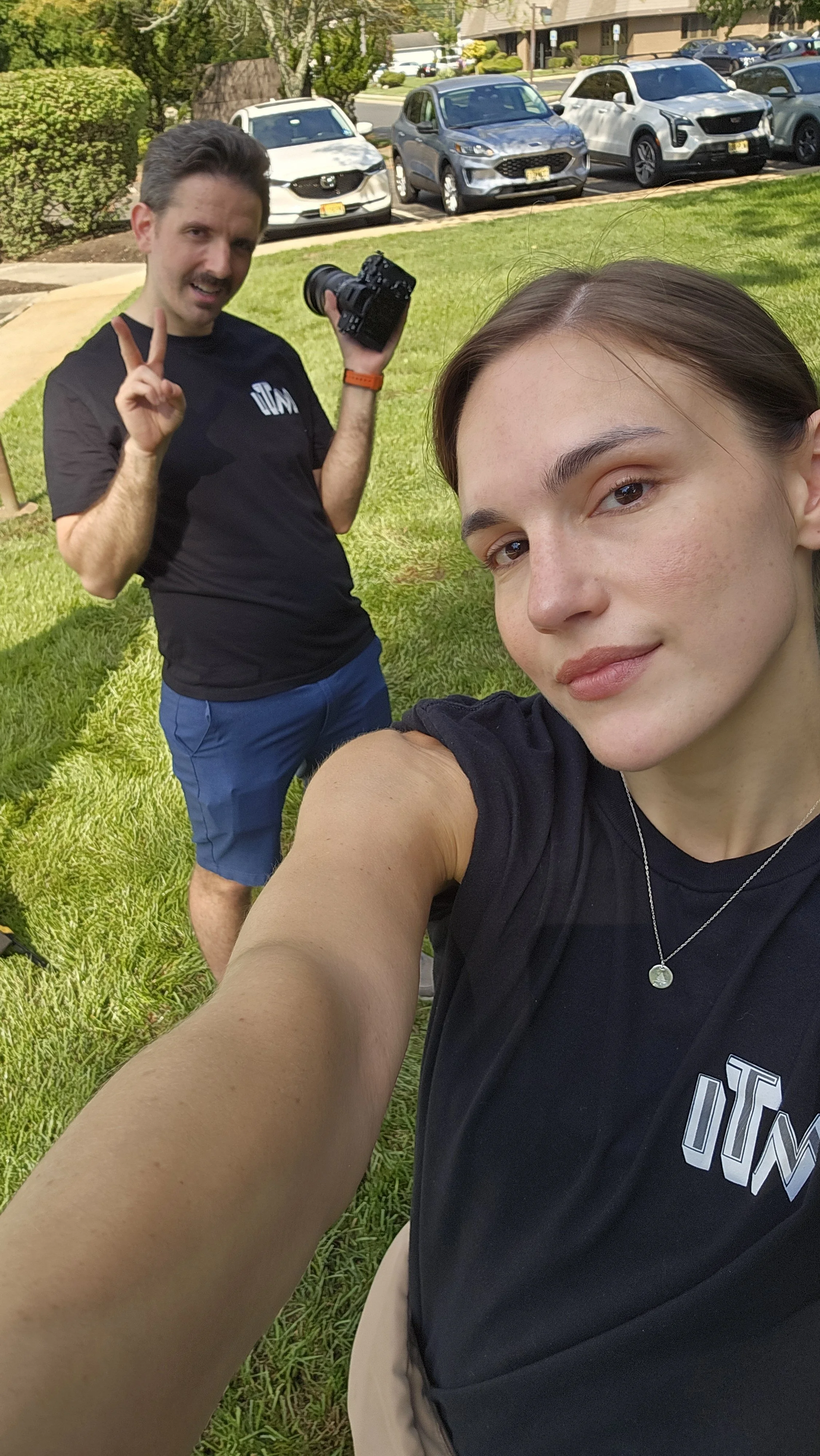 Two young women taking a selfie outdoors on a sunny day with a parking lot and parked cars in the background. One woman is in the foreground, smiling, with long brown hair and wearing a black t-shirt. The other woman is in the background, holding a camera, making a peace sign, with short dark hair and wearing a black t-shirt with a logo and blue shorts.