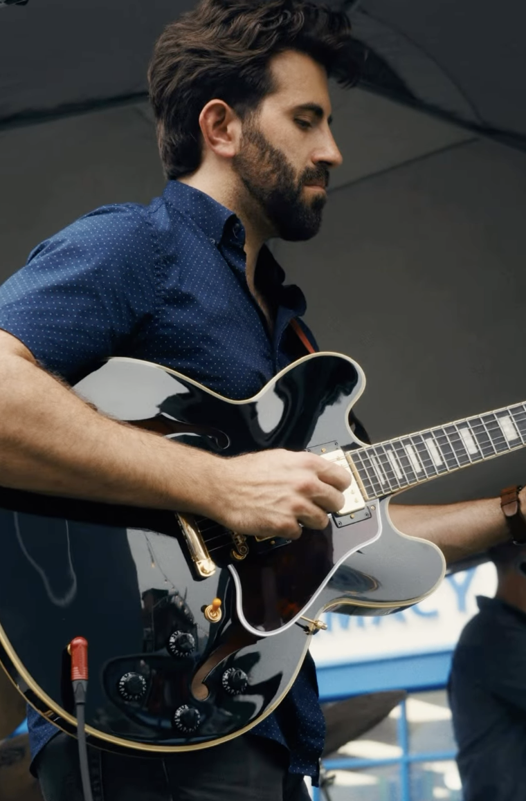 Man with dark hair and beard playing a black electric guitar on stage.
