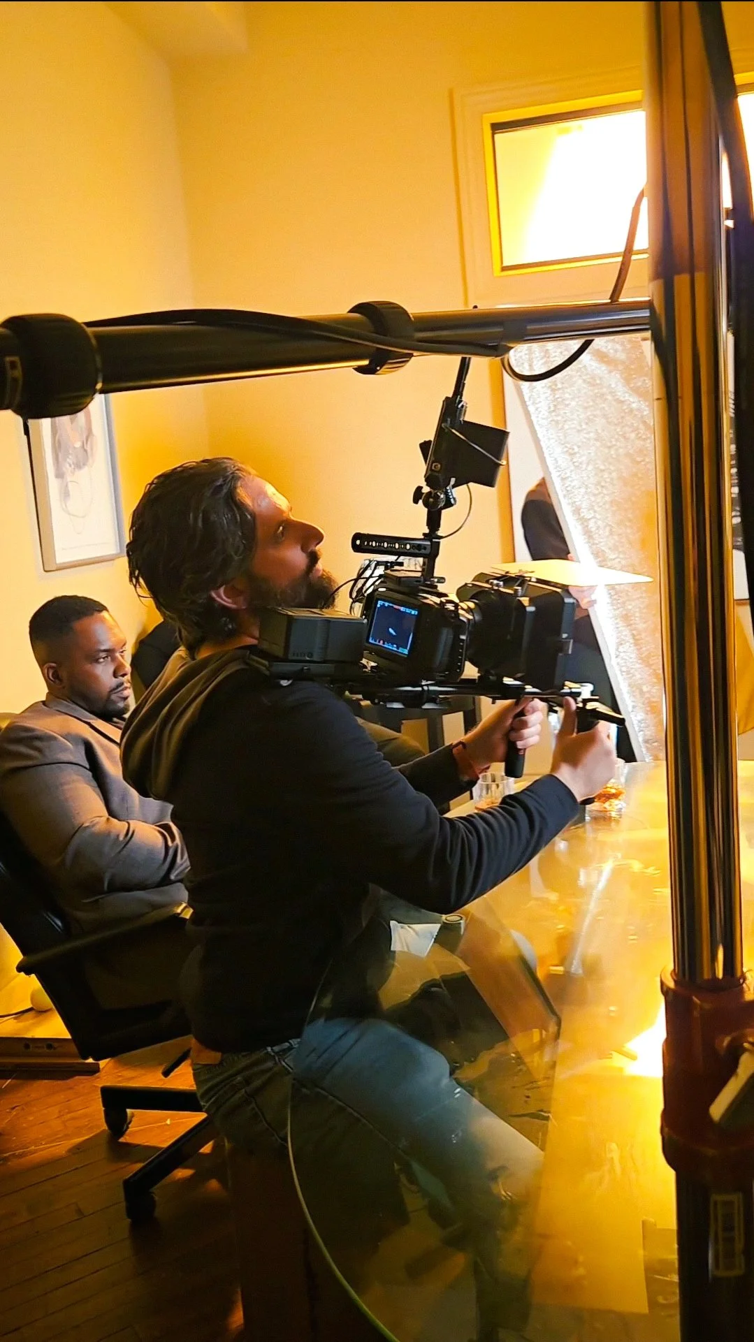 A man operating a professional video camera on a steadicam rig at a conference table during a meeting.