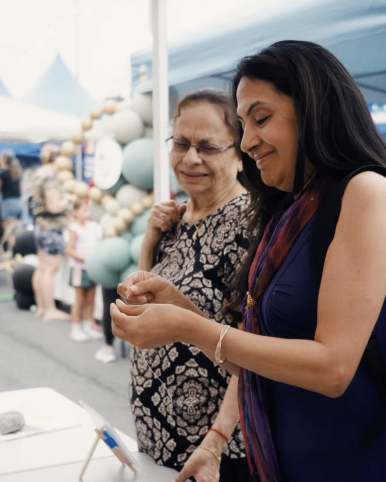 Two women smiling at a photo booth at an outdoor event, with balloons and tents in the background.