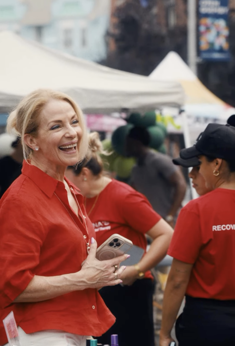 A woman with blonde hair, smiling and holding a smartphone, wearing a red shirt, standing at an outdoor event with tents and other people around.