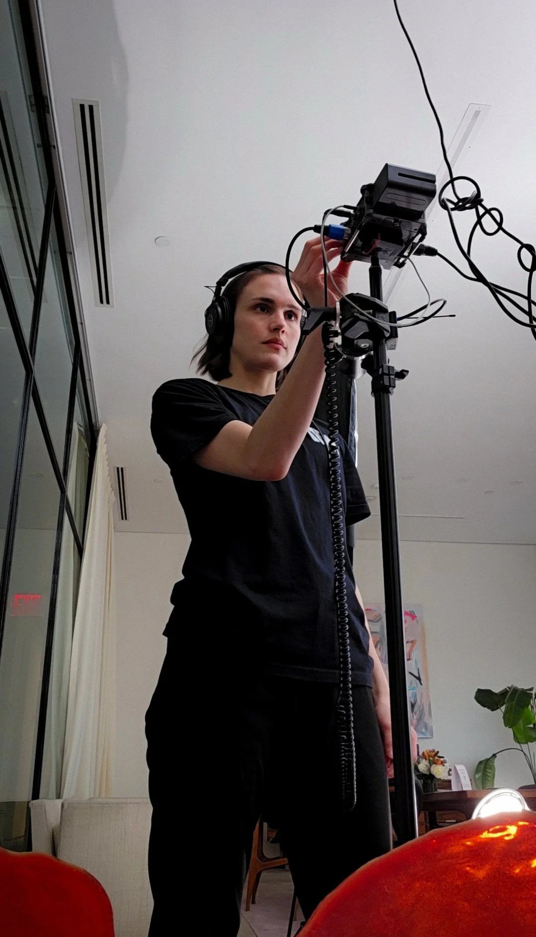 Woman with headphones adjusting equipment on a stand in a room with white walls, curtains, and plants.