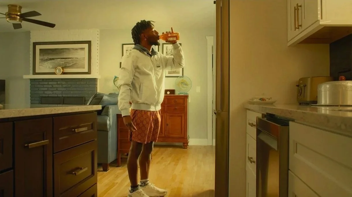 A man standing in a kitchen, wearing orange shorts, a white jacket, drinking from a glass bottle.