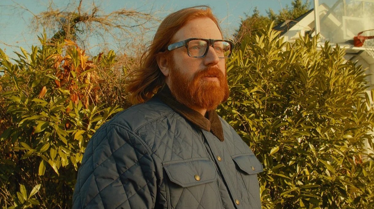 A man with red hair, a beard, and glasses standing outdoors in front of green plants and trees on a sunny day.