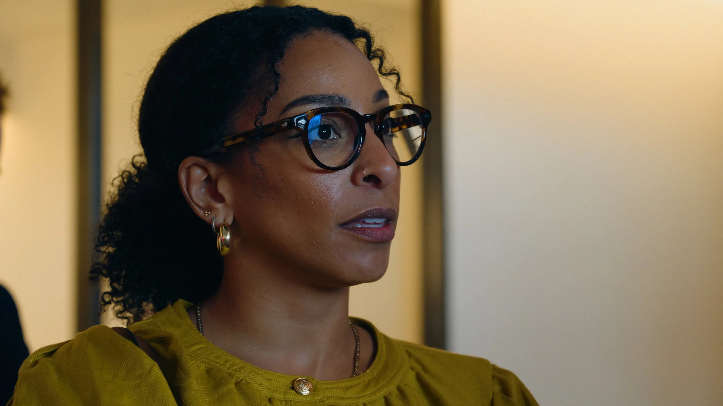 A woman with curly black hair, wearing glasses with tortoiseshell frames and gold earrings, is looking slightly to her left. She is dressed in a yellow top and is in an indoor setting with a blurred background.