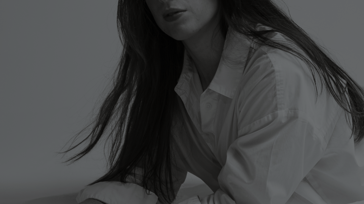 A woman with long dark hair wearing a white collared shirt, leaning forward, dark background.