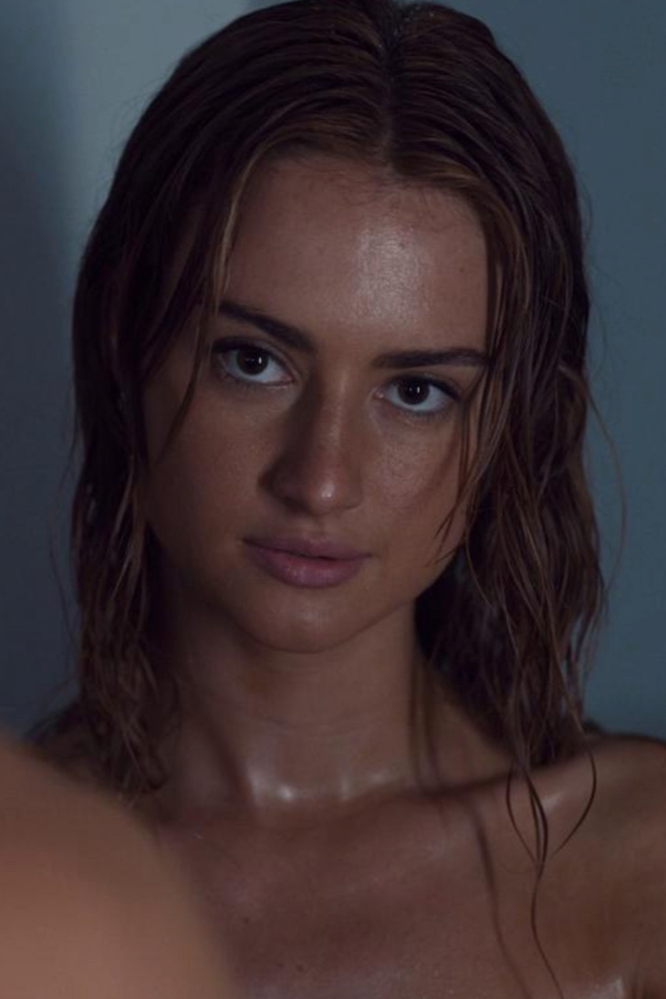 Close-up of a young woman with wet, wavy brown hair, looking directly at the camera with a neutral expression.