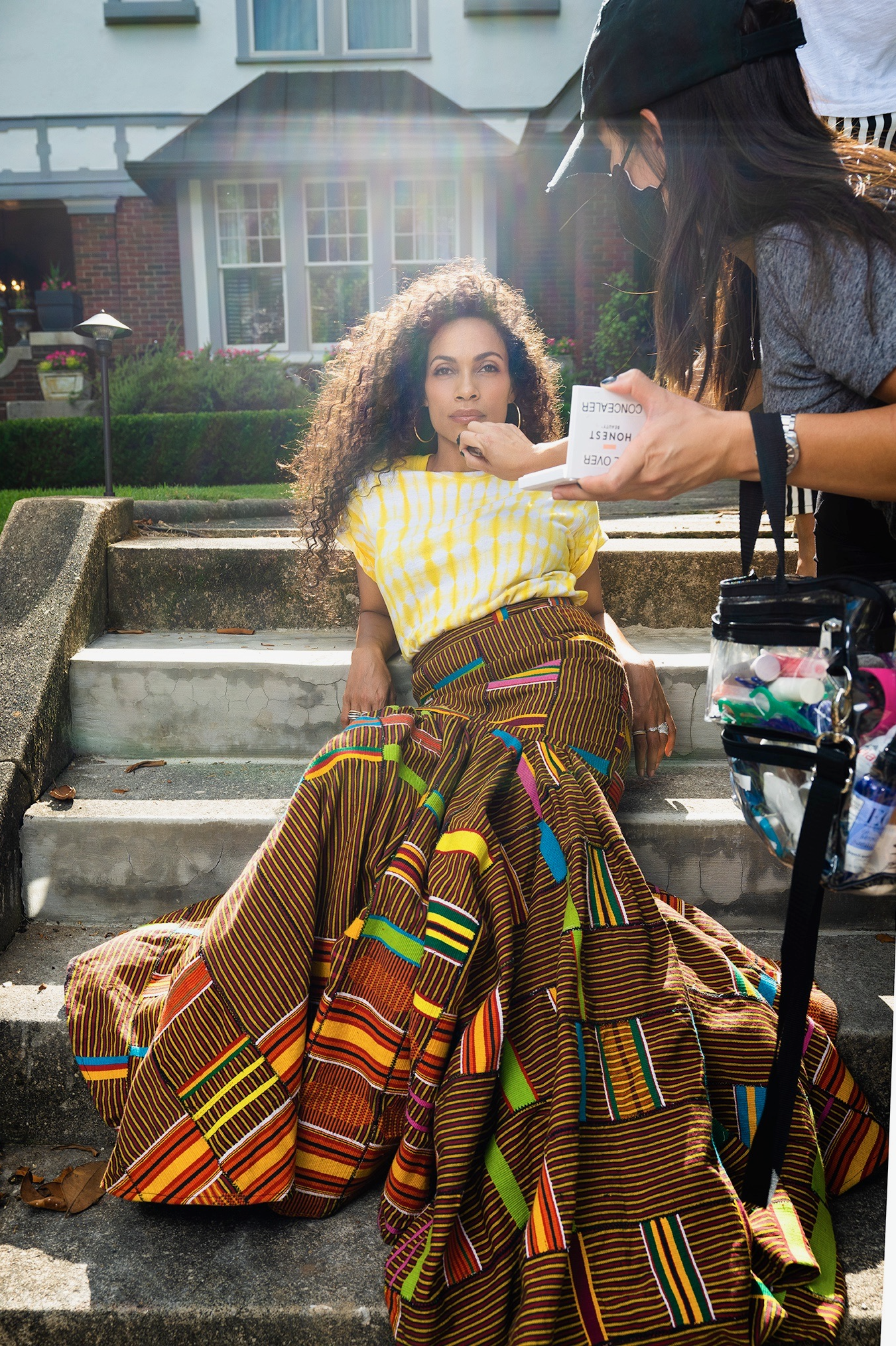 A woman sitting on outdoor stairs being made up by a stylist holding a makeup palette. She is wearing a vibrant, patterned, multicolored skirt and a light yellow top. The setting is outside a residential house with white siding and brick accents, and sunlight is shining from behind, creating lens flare.