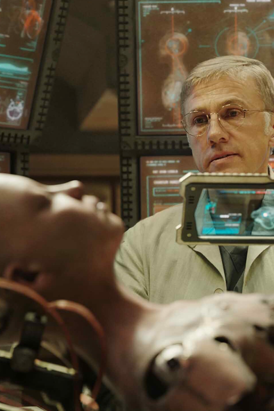 A person lying down connected to medical monitors with electronic wires, while another man wearing glasses and a beige coat observes, with multiple screens displaying scientific data in the background.