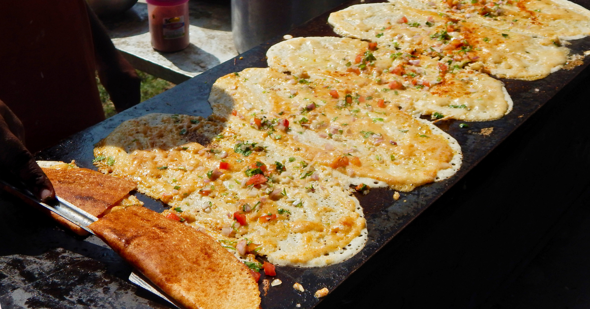 Several dosa crepes cooking on a large flat iron griddle, topped with chopped onion, tomato and fresh coriander