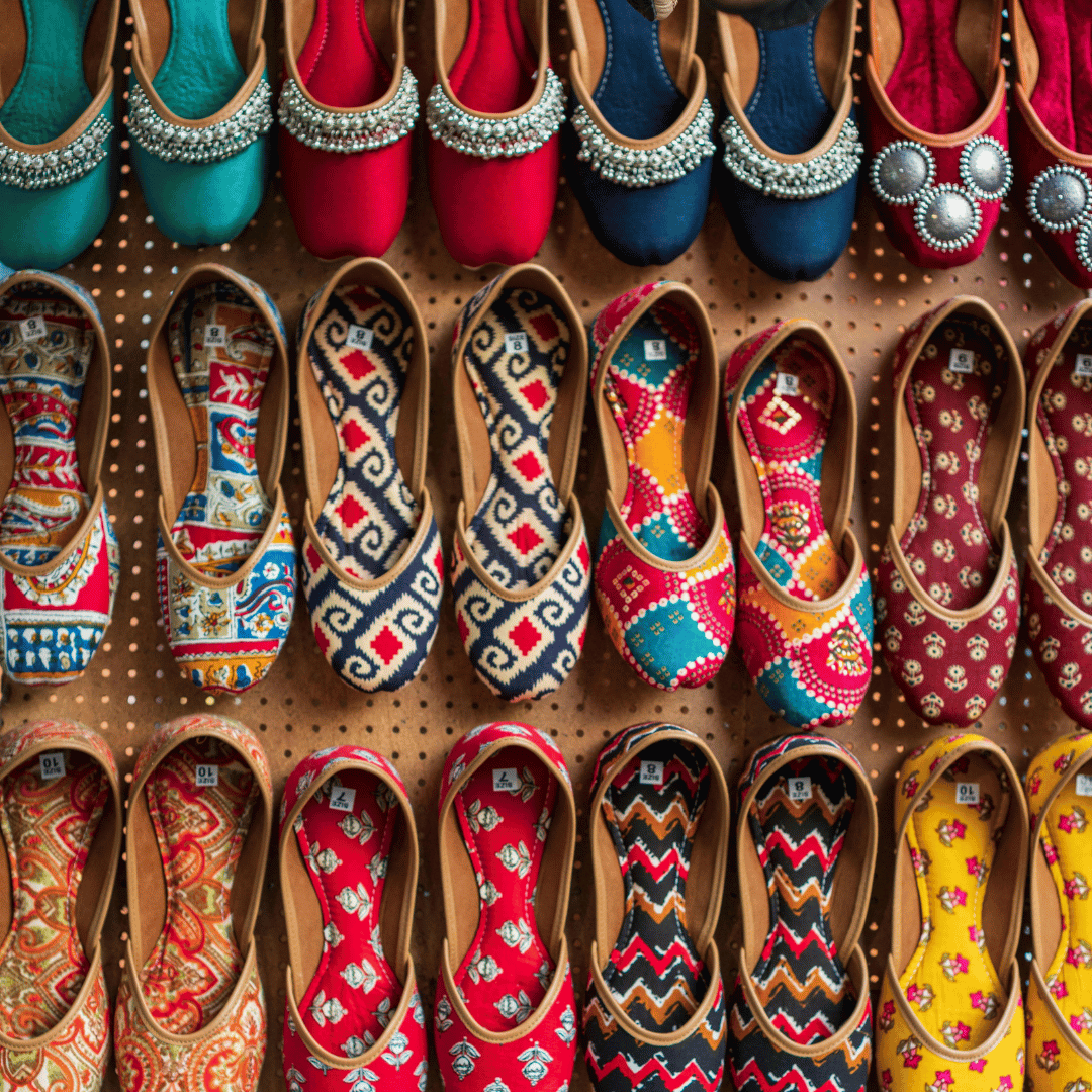 Colorful traditional embroidered shoes displayed on a wall, featuring a variety of patterns and hues like red, blue, and yellow. Some shoes have intricate beadwork.