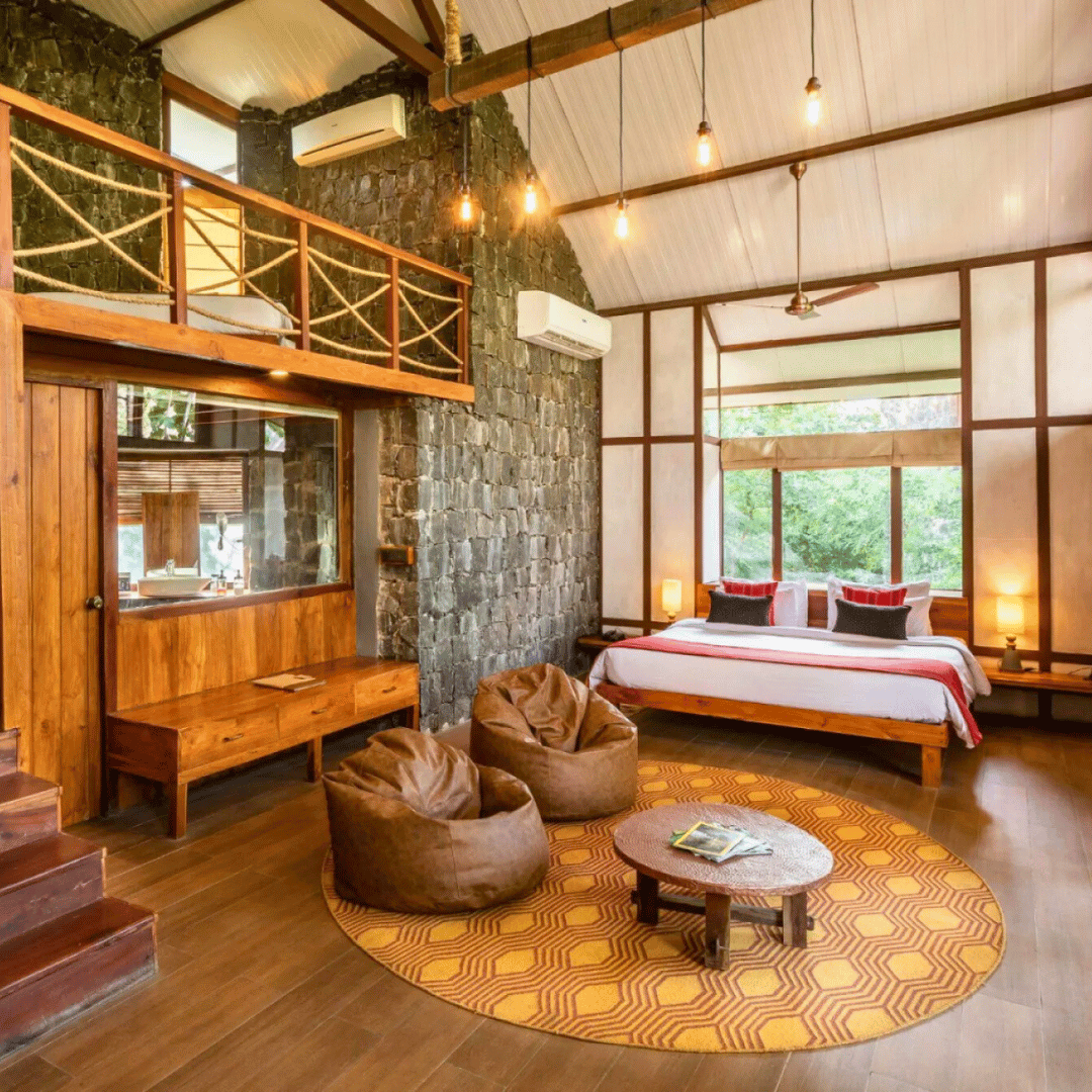 A cozy hotel room with a loft, stone and wood decor, a large bed by windows, two brown bean bags on a yellow patterned rug, and warm lighting.
