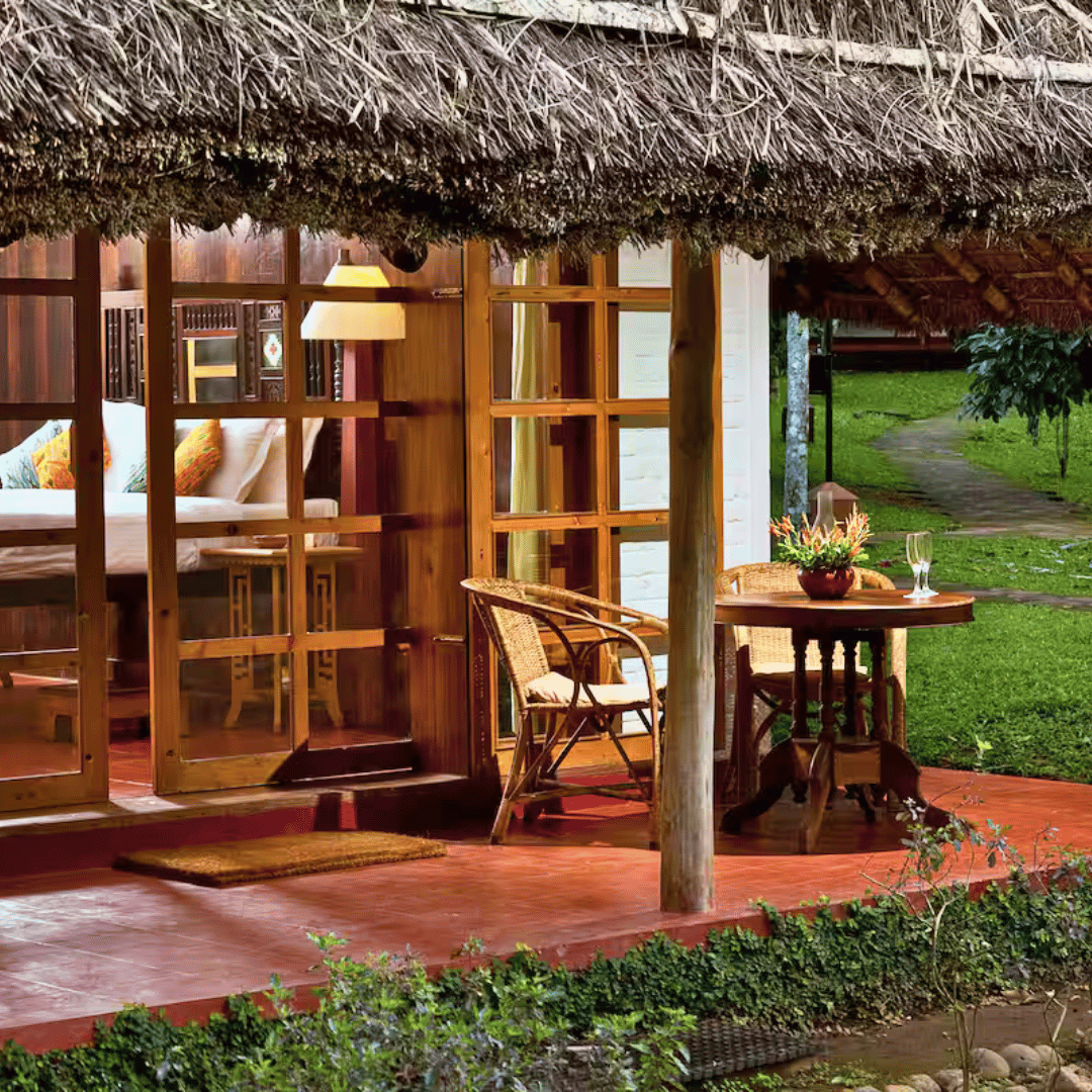 A cozy outdoor patio of a bungalow with a thatched roof, wooden furniture, and a small table with flowers, overlooking a lush green garden and a winding stone pathway.