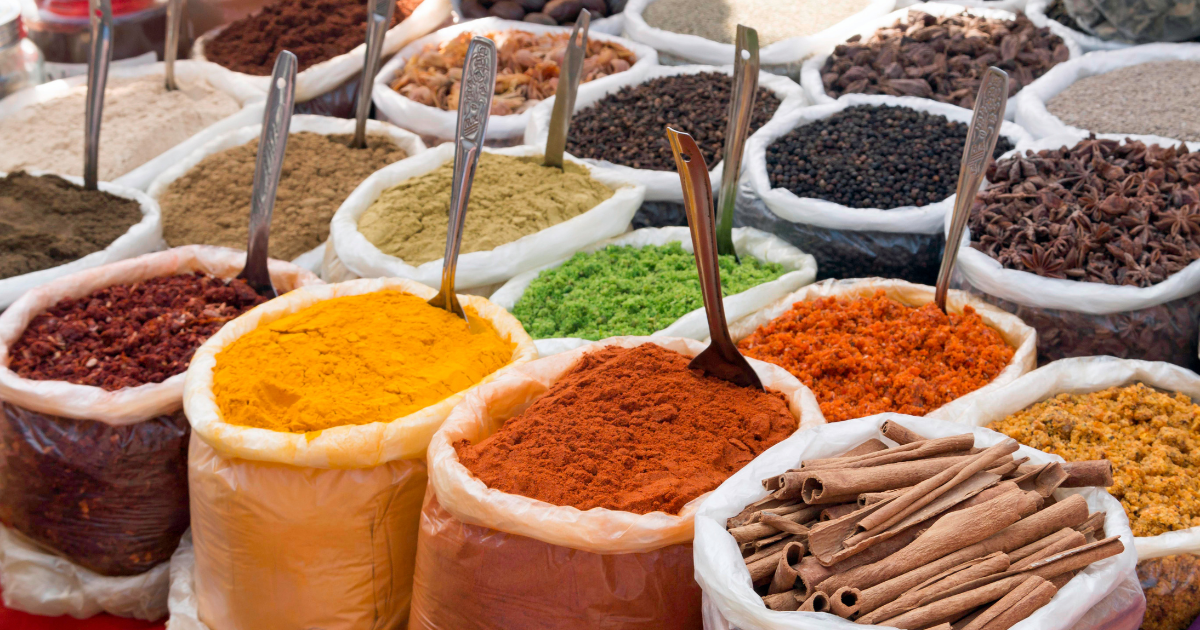 Large sacks of ground and whole spices including turmeric, chilli powder, coriander, star anise and cinnamon displayed at a market stall with metal spoons