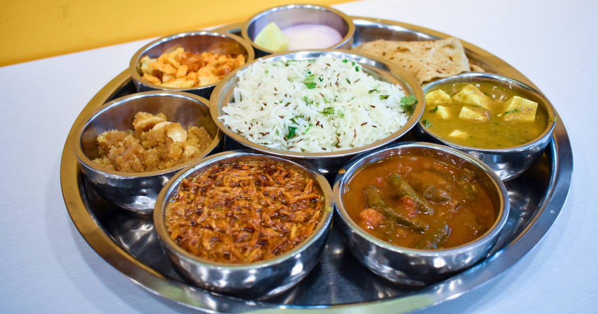 A steel thali plate with small bowls containing jeera rice, two vegetable curries, dal, roti, pickle and raita