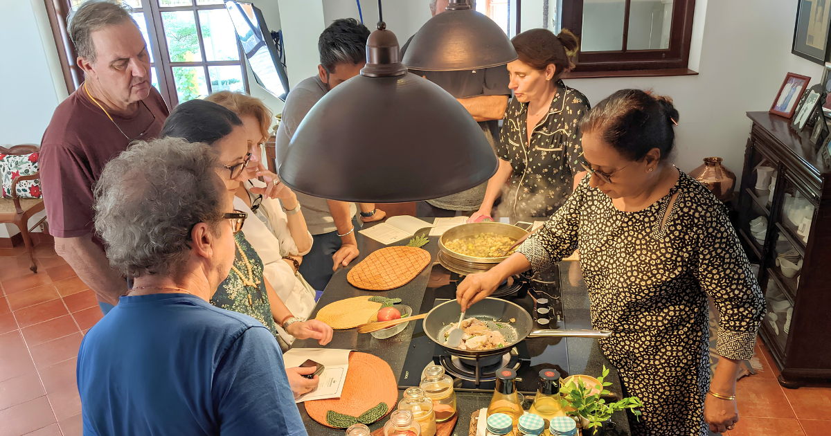 A group of travellers gathered around a kitchen counter watching a local host demonstrate cooking techniques at a home cooking class