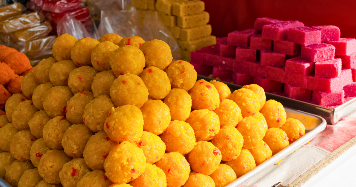 A pyramid of yellow ladoo balls and stacked pink barfi pieces displayed on trays at an Indian sweet shop