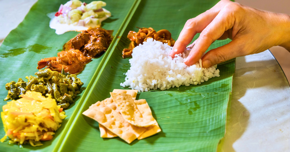 A hand reaches for rice on a banana leaf beside portions of vegetables, curries and a crisp wafer.