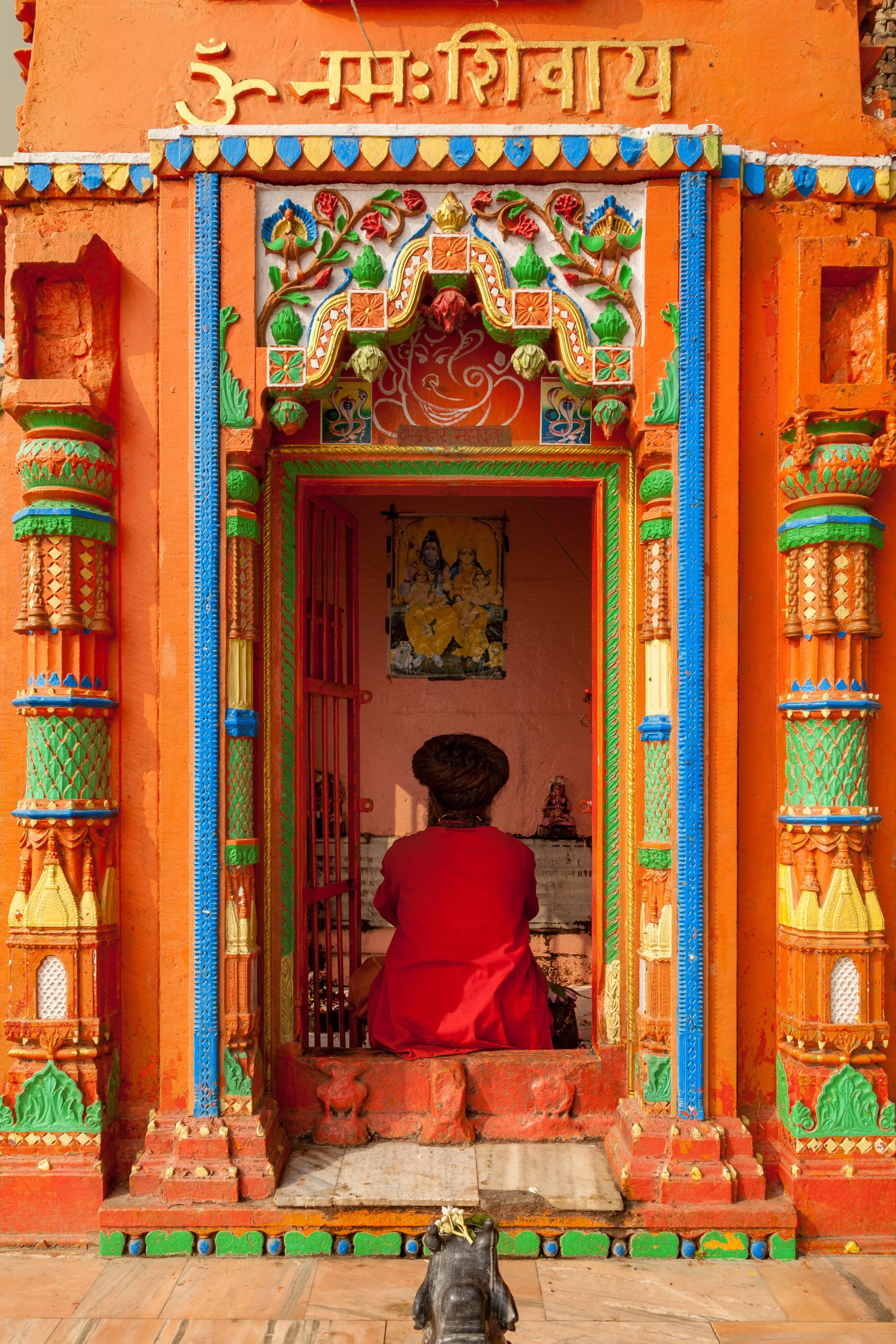A person in a red outfit sitting inside a colorful, ornate Hindu temple entrance with intricate carvings and statues, facing an interior shrine with religious images. A small black animal, possibly a dog, sits outside the temple.