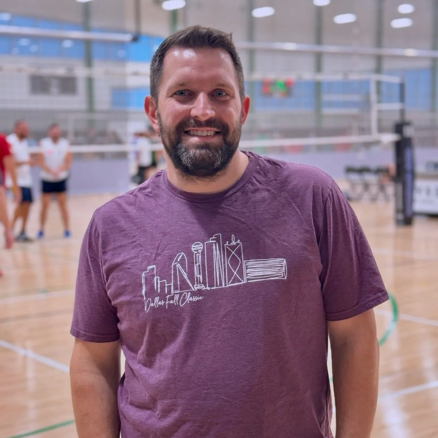 A smiling man with a beard wearing a purple T-shirt standing in an indoor sports gym with volleyball players in the background.